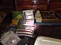 View of multiple pillows including two American flag-themed pillows in plastic packaging and other assorted pillows and blankets beside a red storage box.