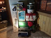 Photo showing stacked coolers (green vintage Coleman, white/red Coleman), red fabric canteen carrier, green Coleman lantern, black tackle box, Emerson insect killer in box, hinged gutter guard box and other items on basement floor.