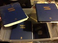 Photo showing multiple blue binders/albums in a plastic container, with white pages inside. Albums labeled with gold lettering and logos.