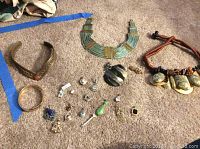 Photo of assortment of costume jewelry including necklaces with natural stone pendants, large green and gold necklace, bangle bracelet, and various small pins and earrings on carpeted floor.