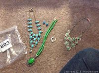 Overview of all jewelry items on carpet: gold-tone blue stone necklace, green bead necklace with pendant, silver-tone green stone cluster necklace, blue stone earrings, and silver-tone ring.