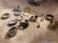 Photo showing the full collection of 14 bracelets including cuffs and bangles in silver and gold tones, some with stones and metal detailing, laid out on carpeted floor.