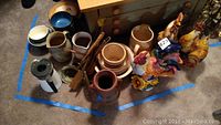 Photo showing a collection of ceramic bowls, pitchers, kitchen utensils, and colorful ceramic roosters arranged on the floor.
