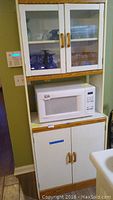Front view of white storage cabinet showing upper glass doors with wooden handles, middle microwave shelf with microwave, and lower cabinet doors.