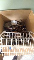 Box containing stacked copper pans and metal cooking vessels with a white metal door rack in front