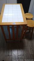 Kitchen table showing rectangular top with white ceramic tiles and wooden frame, with two round wooden stools nearby.