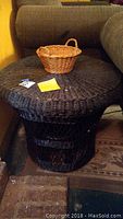 Side view of one black wicker table with a small wicker basket on top, placed next to a brown couch and over a wooden floor.