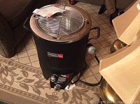 Front view of black infrared turkey fryer with stainless steel mesh basket and lid sitting on carpet.