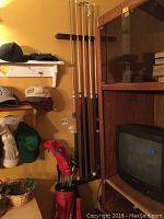 Photo showing four wooden pool sticks mounted vertically on the wall beside a cabinet and some hanging hats.