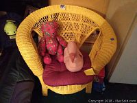 Front view of yellow wicker chair with red cushion and two plush toys: pink teddy bear and pink swan.