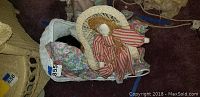 Photo of rag doll seated in the white wicker pram on a carpeted floor, showing the doll dressed in a red and white striped dress with a bonnet and the pram's woven wicker design.