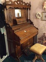 Front angled view showing ornate carvings, mirror, and keyboard cover of antique organ with upholstered stool in front.