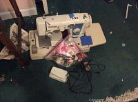 Full view of the Morse sewing machine on a blue carpeted floor next to a wooden table leg, with foot pedal and bag of sewing notions in front.