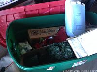 Photo of green plastic tote containing various Christmas decorations including mini artificial trees, teddy bear, red pillows, and cardboard box