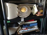 Shelf with two fans, small mirror with white frame, brown teddy bear, and multi-colored box on top shelf. Board games and white storage or bag holders on lower shelf.