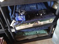 Camping lot items stacked on shelving showing two air compressors, a blue bag with Columbia branding, tent carrying bag, and rolled floor mats.