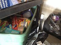 Photo showing a green plastic bin filled with various kitchen wraps and storage items, including boxes of plastic wrap, aluminum foil, and dust masks, along with two rolls of tape and a plastic container with blue lid.
