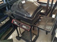 Full view of black barbecue grill with dome lid, side shelves, two wheels, and signs of wear. Missing one leg and holder.