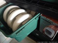 Five white oscillating fans stored in a large green plastic bin and a boxed NOMA ceramic heater next to the bin.