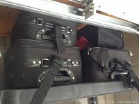 Photo showing two large black fabric suitcases with buckled straps and handles stacked side by side inside an open compartment.