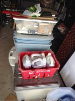 Stack of plastic totes including clear, blue, and gray totes with a red crate on top containing various automotive fluid containers and cleaning brushes visible on top.