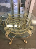 Round glass table with clear tempered glass top and distressed white metal base with scrollwork.