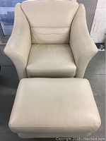 Front view of beige leather armchair with matching ottoman in front of it on concrete floor.