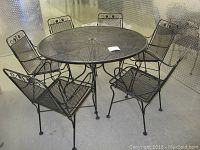 Front view of round wrought iron patio table and six chairs showing mesh table top and chairs with leaf themed backs.