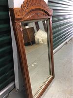 Full view of the wood framed wall mirror leaning against a metal storage unit with concrete floor. Mirror size approximately 19.5 by 35 inches.