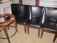Four black upholstered dining chairs with wood legs, placed in a row next to a round table base missing its glass top, showing wear and scratches