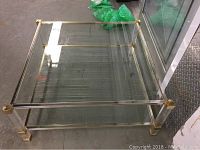 Angle view of the glass top coffee table showing the clear glass top and brass-colored frame with acrylic legs.