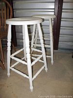 Front and side view of two white wooden stools with turned legs showing paint chips and wear, placed on concrete floor.