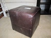 Black leather-like cube ottoman with visible wear and stitching, placed on concrete floor.