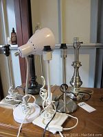 Seven table lamps arranged on a table showing various metal finishes, round and square bases, no lampshades, white cords coiled around bases