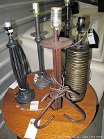 Five different table lamps displayed on a wooden table. The bases vary in design including spiraled, ridged, rusted metal, coiled bronze, and columnar shapes.