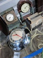 Three items on a glass table: wood folding mantel clock, chrome Kelton electric wall clock, and boxed Agfa Ansco camera.