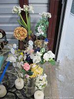Overview showing assorted faux flowers including white lilies, brown hydrangea-type flower, white orchids with other mixed colors, and a dried flower arrangement in vases on a glass table with cream candles nearby.