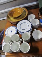 Lot of stoneware plates, bowls, cups, and saucers arranged on wooden table, showing four large golden-yellow Netas plates, blue and white floral small plates, and multiple cups and saucers with blue rims.
