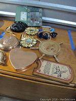 View of copper colored fish wall art, copper colored fish pan, heart-shaped copper pan, two small decorative mirrors, black masquerade mask, wooden boat steering wheel missing a piece, plaque from Argentina, and a small cup holder from Cuba arranged on wooden surface.