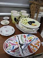 Eight rooster plates stacked with four matching rooster mugs behind them, small floral plate and Japanese small plate at the side, plus white rack stand beneath