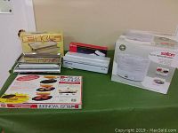 Full view of all kitchen items on green table including EKS scale with box, food dehydrator box, food sealer, and polythene rolls