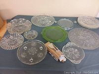 Wide view of the lot showing various clear glass plates and bowls, plus green depression glass plate and wooden-handled knives.