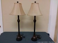 Photo of pair of brown table lamps with fabric bell-shaped shades on a blue surface against a beige wall.