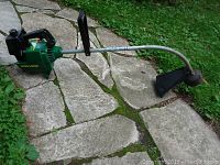 Full view of the green Weed Eater gas trimmer model GTI 15 with curved shaft and handle, resting on a stone walkway with grass around.