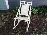 Front and side view of small white wooden rocking chair with woven backrest on garden soil.