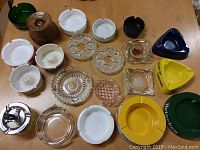 Overhead view of assorted ashtrays on a wooden table showing a variety of colors, materials, and styles including clear glass, ceramic, metal, and wood ashtrays.