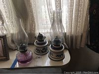 Three oil lamps arranged on a surface in front of lace curtains. Includes two Pennsylvania Dutch style lamps and one cut glass lamp with purple base.