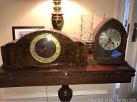 Wide shot showing both mantle clocks on a wooden surface with items in the background including a lamp and model ship.