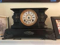Front view of black antique mantle clock with gold bezel and Roman numeral dial, missing glass, with winding keys on shelf below.
