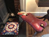 Photo showing embroidered top stool with a clock and teacup design, wooden bucket, leather topped stool and some fireplace tools near a fireplace
